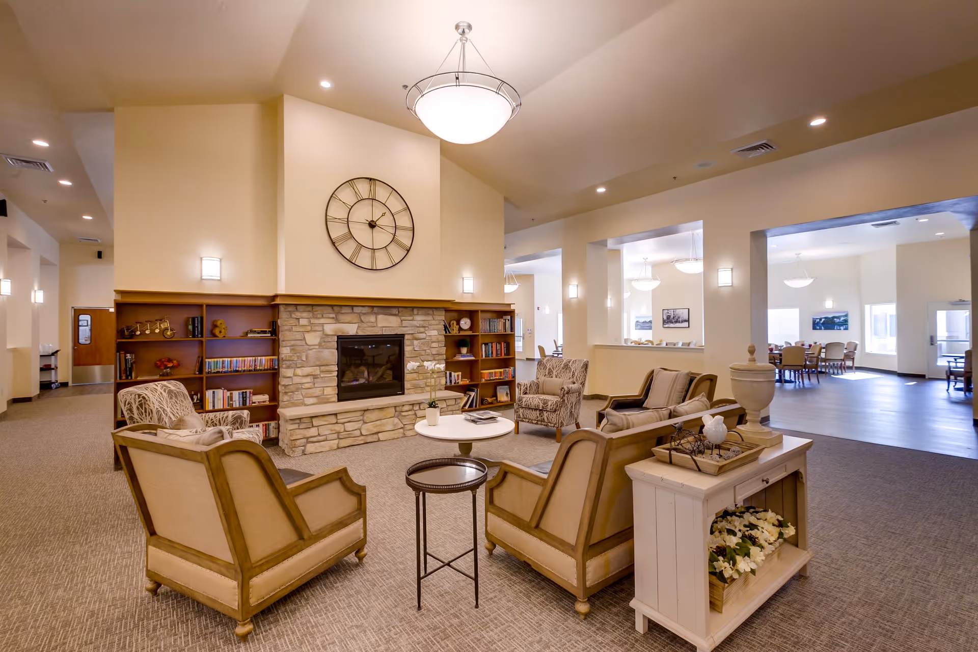 A cozy senior living common area featuring a stone fireplace with a large clock above it, surrounded by built-in wooden bookshelves. The seating area includes two beige armchairs and two patterned armchairs arranged around a small round table. The room is well-lit with ceiling lights and wall sconces, and there is a white console table with decorative items and flowers. In the background, a dining area with tables and chairs is visible through large open wall sections.