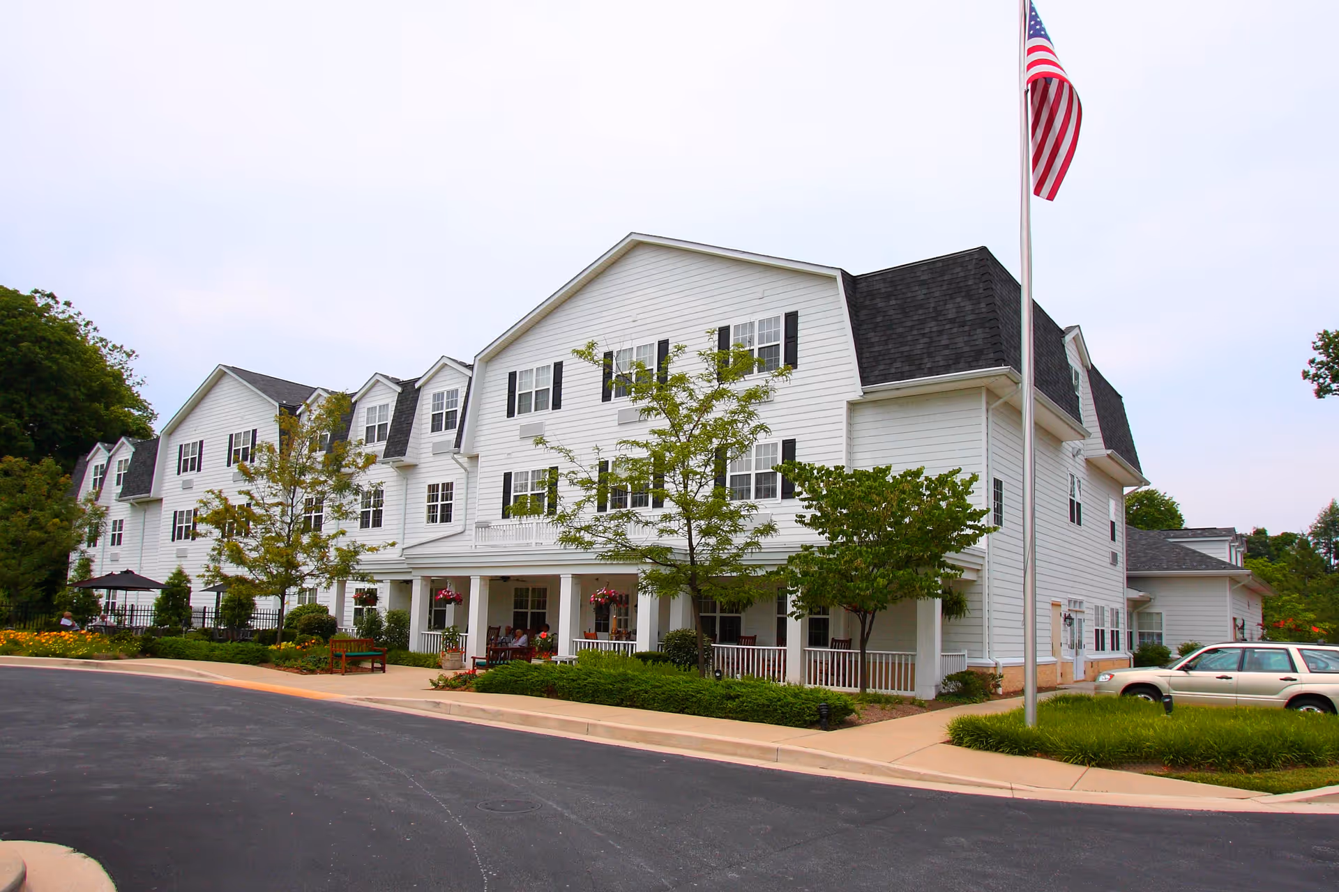White three-story senior living building with a front porch, landscaped grounds, parked car and an American flag.