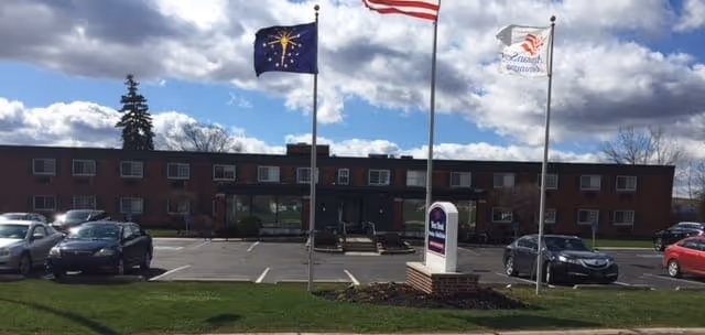 Front exterior view of West Bend Nursing and Rehabilitation building with three flagpoles displaying the American flag, the Indiana state flag, and a facility flag. Several cars are parked in the parking lot in front of the building under a partly cloudy sky.