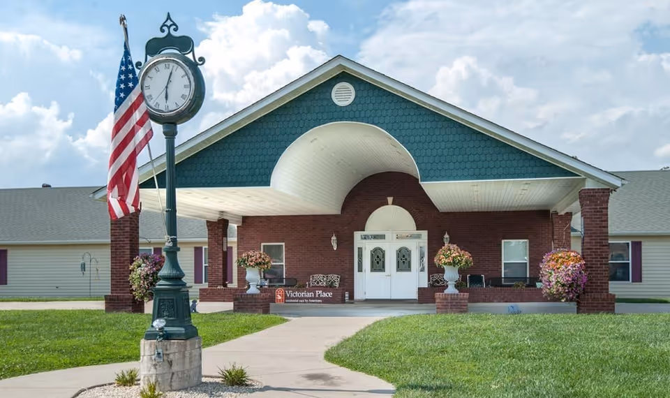 Exterior view of Victorian Place of Owensville, brick entrance, clock, flag, and flower planters.