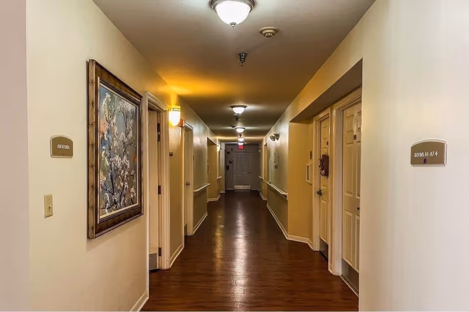 A well-lit hallway in a senior living facility with wooden flooring and cream-colored walls. Several doors line both sides of the corridor, with signs indicating room numbers and an emergency exit. A framed floral painting is hung on the left wall.
