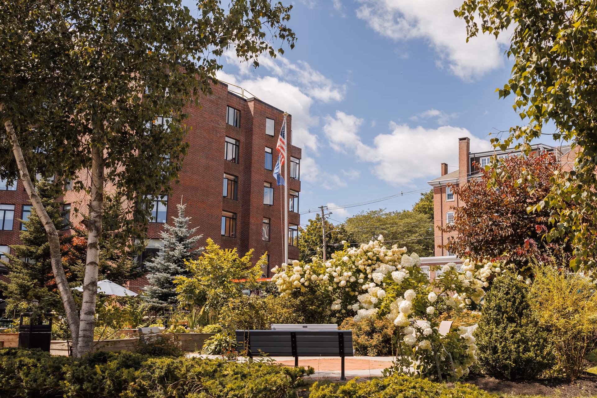Outdoor garden area with a variety of trees, bushes, and blooming white flowers. There is a black bench in the foreground and two brick buildings in the background under a partly cloudy blue sky. Two flagpoles with flags are visible near the center of the image.
