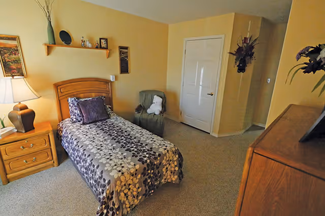 A cozy bedroom with a single bed covered in a floral patterned bedspread in shades of purple and beige. There is a wooden nightstand with a lamp and a book on it to the left of the bed. Above the bed, a wooden shelf holds decorative items and framed pictures. A green armchair with a white teddy bear sits in the corner near a closed white door. The walls are painted a warm beige color, and there is a wooden dresser on the right side of the room with a vase of flowers on top.