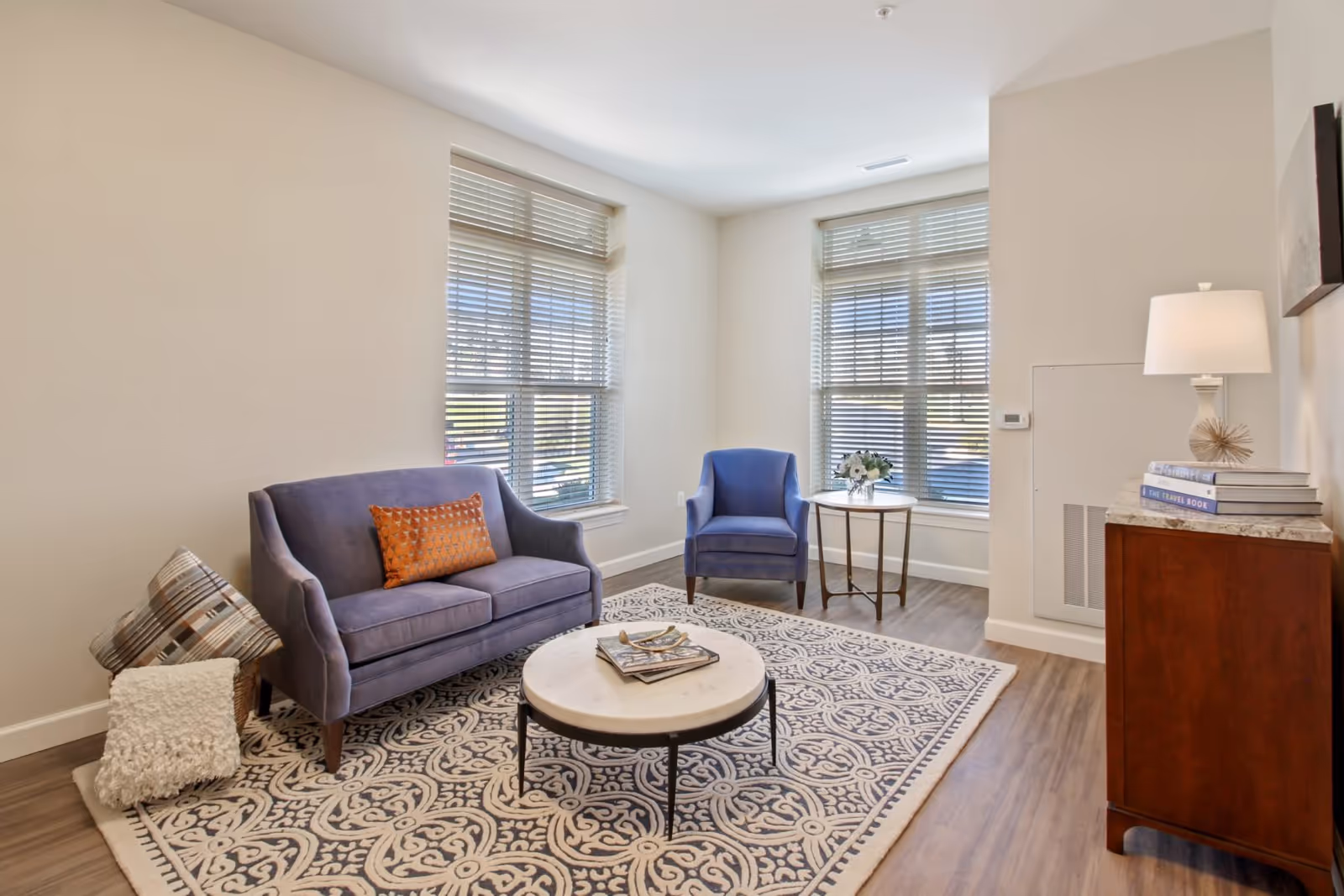A bright and cozy living room with two large windows covered by blinds, a purple loveseat with an orange pillow, a matching purple armchair, a round coffee table with books and decorative items, a patterned area rug, and a wooden cabinet with a lamp and stacked books.