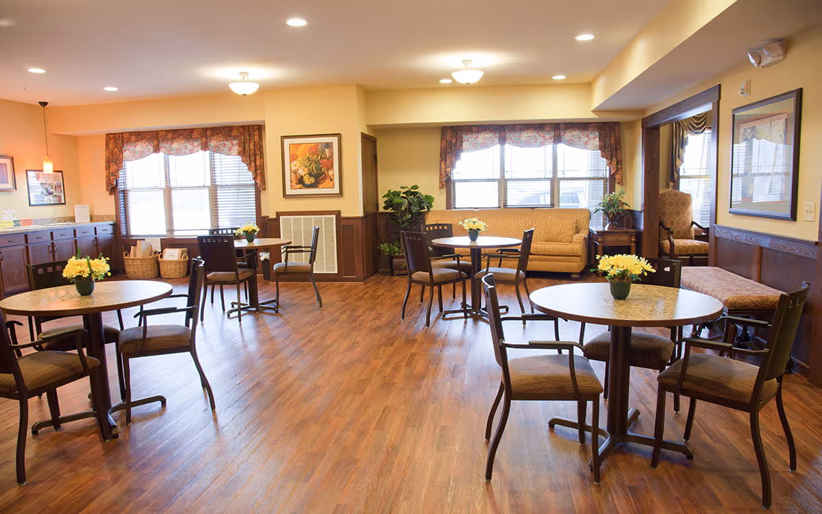 Spacious senior living common room with round dining tables and chairs, a sofa, decorative plants, and wood flooring.