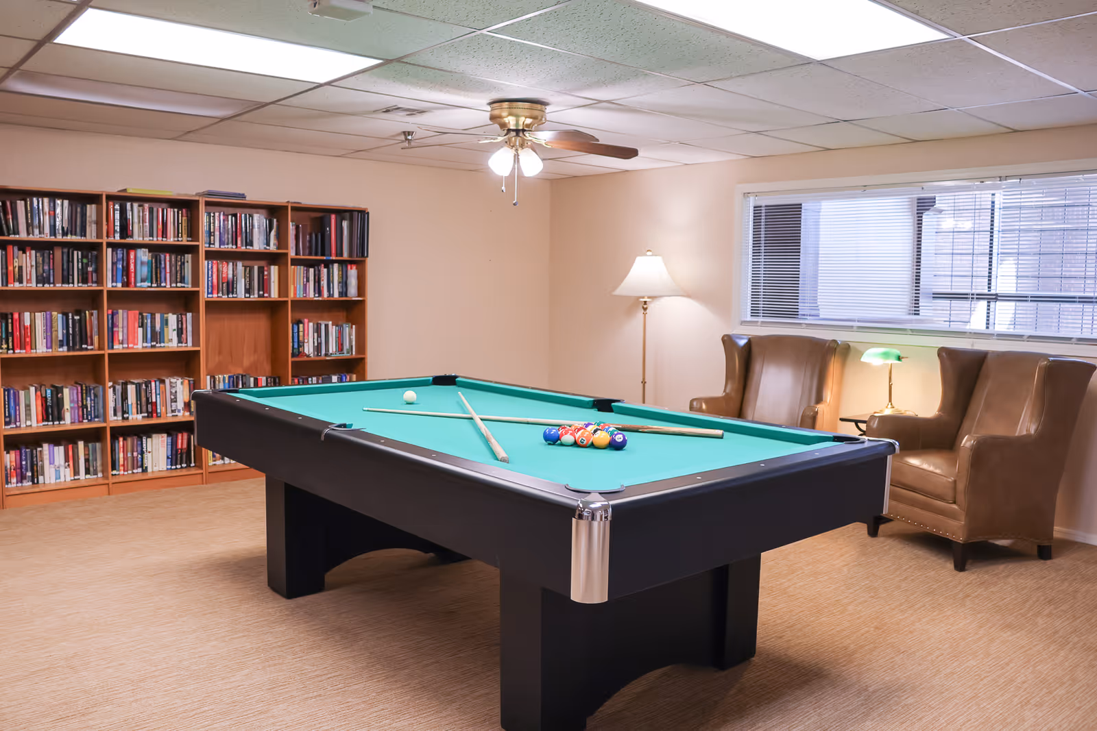 A recreational room with a pool table in the center, two brown leather armchairs near a window with blinds, a floor lamp, a small table with a green desk lamp, and bookshelves filled with books along the wall.