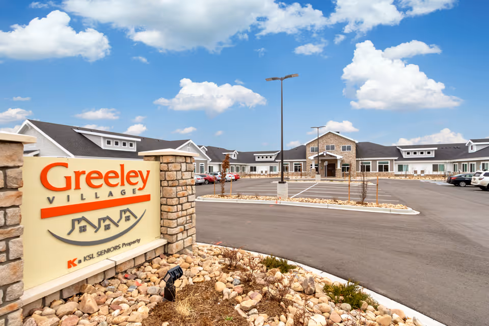 Exterior view of Greeley Village senior living facility with a large parking lot, stone and brick building facade, and a clear blue sky with scattered clouds. A sign with the name 'Greeley Village' is visible in the foreground surrounded by rocks and landscaping.