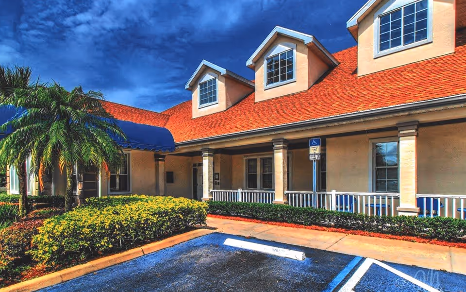 Exterior view of a building with a red roof and beige walls, featuring three dormer windows. There is a blue awning over the entrance, palm trees, and neatly trimmed bushes in front. A handicapped parking space is visible in the foreground under a bright blue sky with some clouds.