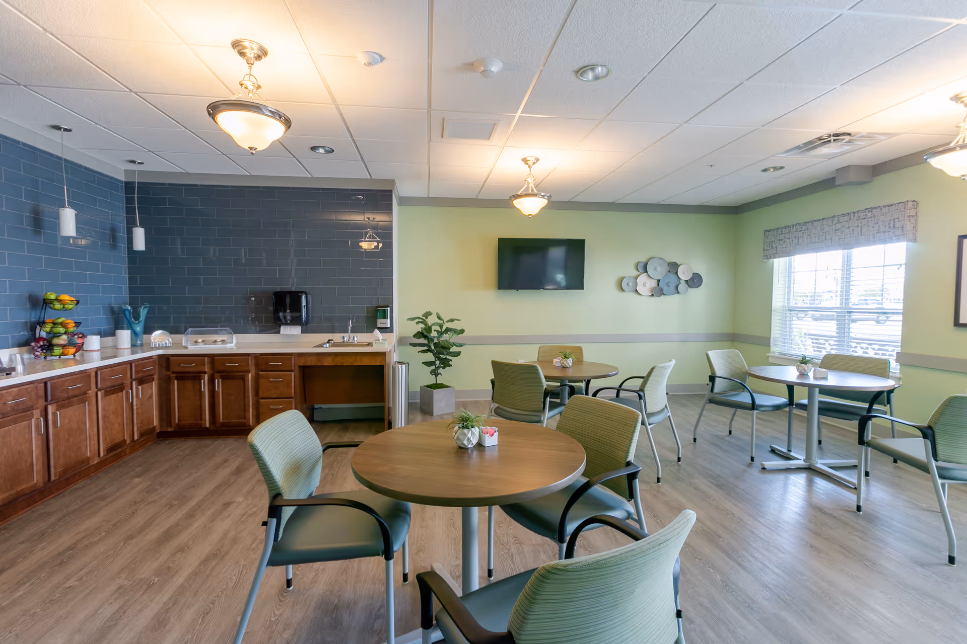A bright dining area with round wooden tables and green cushioned chairs. The room has light green walls, a large window with blinds, and a wall-mounted TV. To the left, there is a kitchen counter with wooden cabinets, a sink, and a blue tiled backsplash. The floor is wood-style laminate, and there are ceiling lights providing illumination.