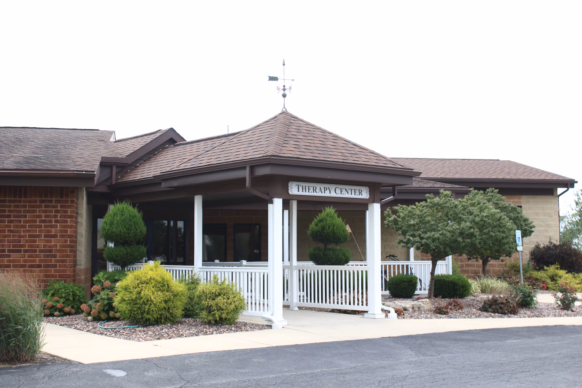 Exterior view of the Bethesda Care Center Therapy Center entrance with a covered porch supported by white columns, surrounded by landscaped bushes and trees.