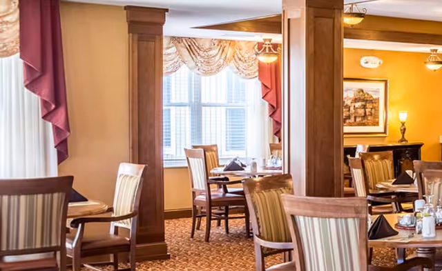 A warm and inviting dining room with multiple wooden tables and chairs arranged neatly. The room features large windows with decorative curtains, soft yellow walls, and wooden columns. Tables are set with napkins and condiments, creating a cozy atmosphere.