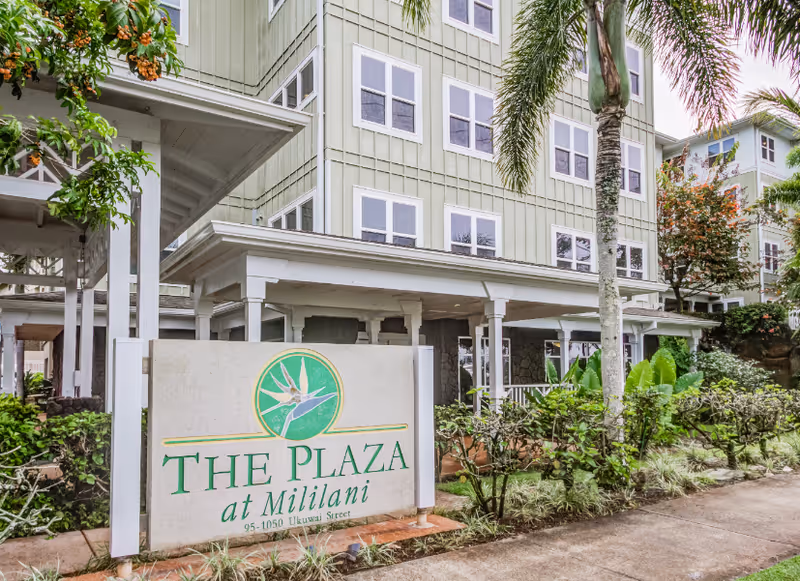 Exterior view of The Plaza at Mililani senior living facility showing a multi-story building with light green siding, multiple windows, palm trees, and a sign with the facility name and address in front surrounded by greenery.