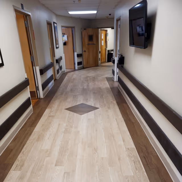 A clean, well-lit hallway in a senior living or rehabilitation facility with light wood flooring featuring a darker diamond pattern in the center. The hallway has beige walls with dark brown handrails on both sides, several open doors leading to rooms, and a wall-mounted television on the right side.