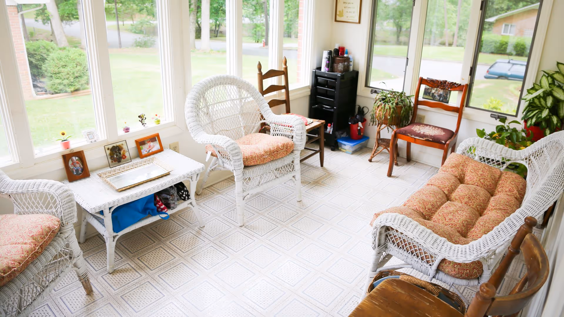 A bright sunroom with large windows showing a green outdoor view. The room contains white wicker furniture with patterned cushions, including chairs and a loveseat. There is a white wicker table with framed photos and small decorative items on it. Additional wooden chairs and potted plants are placed around the room, creating a cozy and inviting atmosphere.