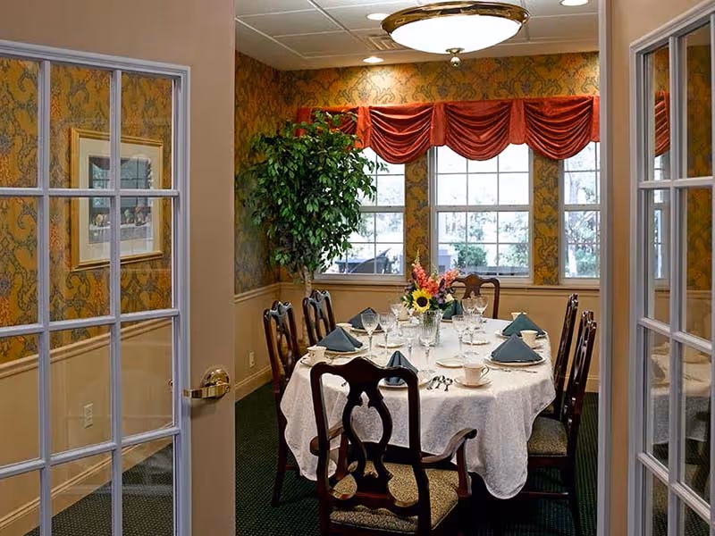A formal dining room with an oval table set for eight, wooden chairs, a floral centerpiece, and windows with red valances.