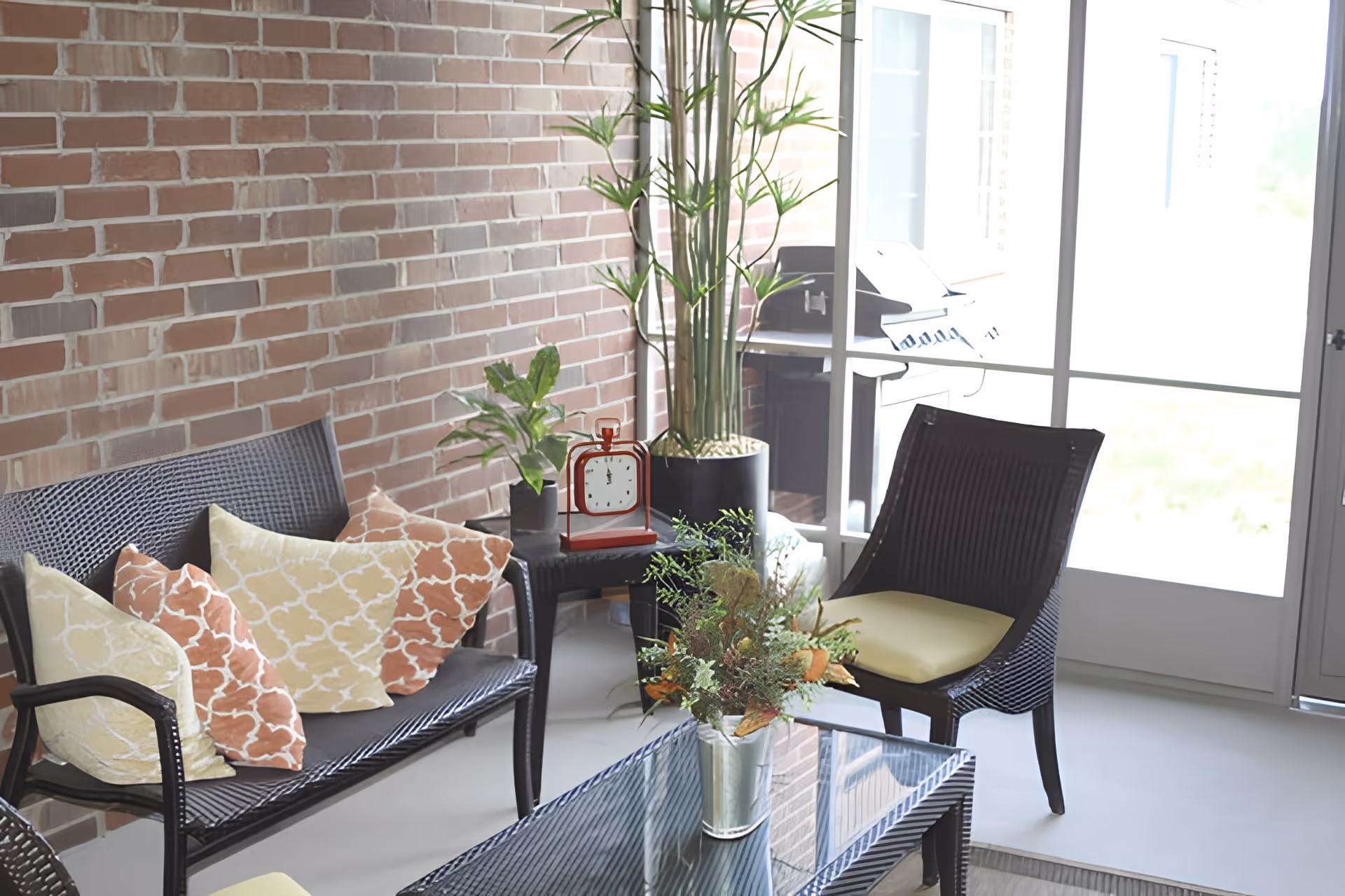 Sunlit seating area with wicker loveseat and chairs, patterned pillows, plants, a glass coffee table and a red clock against an exposed brick wall.