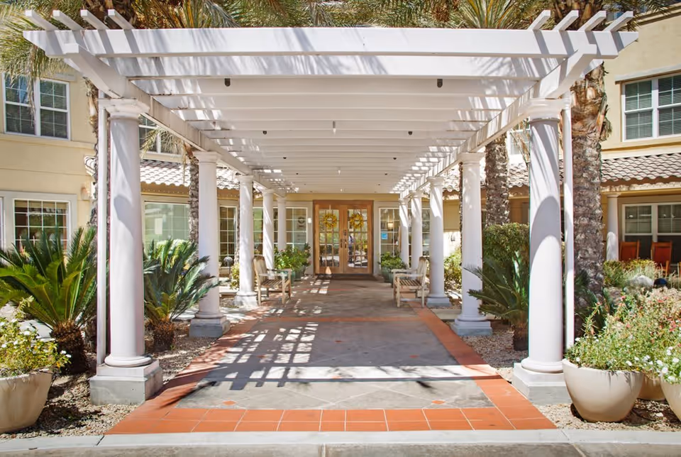 Entrance walkway to a senior living facility with white pergola supported by columns, potted plants, benches, and palm trees on either side leading to double glass doors decorated with wreaths.