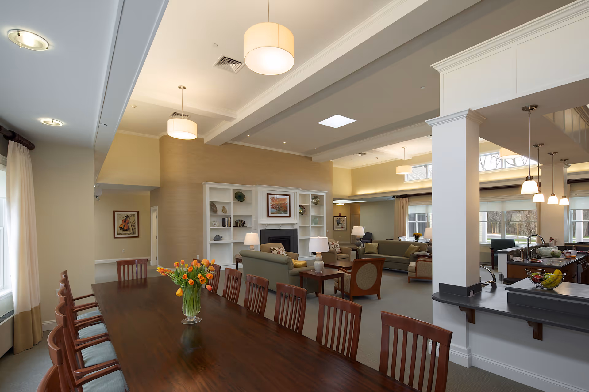 Spacious and well-lit common area in a senior living facility featuring a long wooden dining table with chairs and a vase of orange tulips, a cozy living room with sofas and armchairs around a fireplace with built-in shelves, and a kitchen area with a countertop and hanging pendant lights.