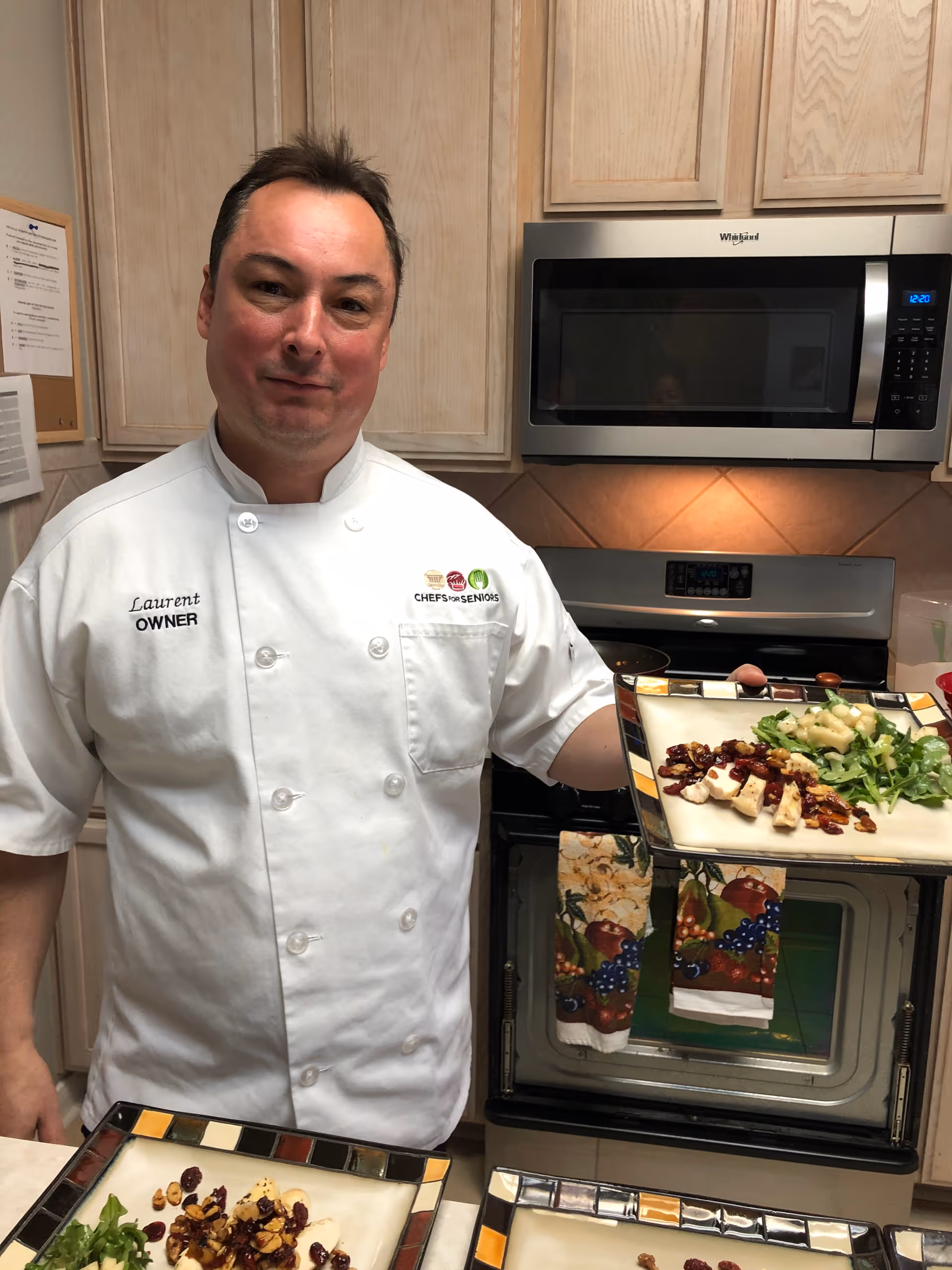 A man wearing a white chef's coat with the name Laurent and title Owner embroidered on it stands in a kitchen holding a plate of food. The kitchen has light wood cabinets, a microwave, and an oven with a colorful towel hanging on its handle. There are additional plates with food on the counter in front of him.