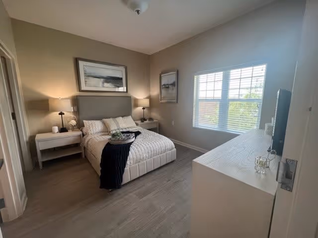 A neatly arranged bedroom with a bed centered against the wall, flanked by two nightstands each with a lamp. The bed is made with light-colored bedding and a dark throw blanket. Above the bed hangs a large framed abstract artwork. There is a window with blinds on the right wall letting in natural light. A dresser with a TV on top is positioned opposite the bed. The room has light-colored walls and flooring, creating a bright and cozy atmosphere.