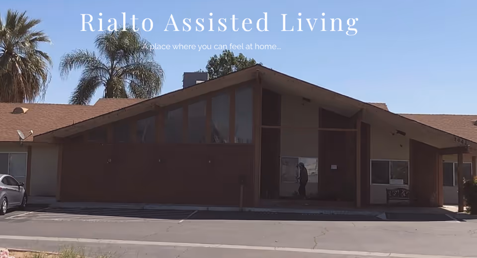 Front exterior of the Rialto Assisted Living building with a peaked roof, palm trees behind, and a parking area in front.