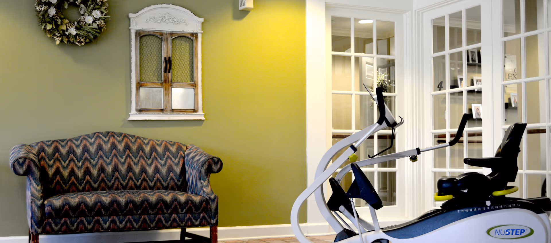 A cozy interior space with a patterned upholstered loveseat against a green wall, decorated with a wreath and a small vintage-style cabinet. In front of the loveseat is a NuStep exercise machine, and behind it are white-framed glass doors leading to another room.