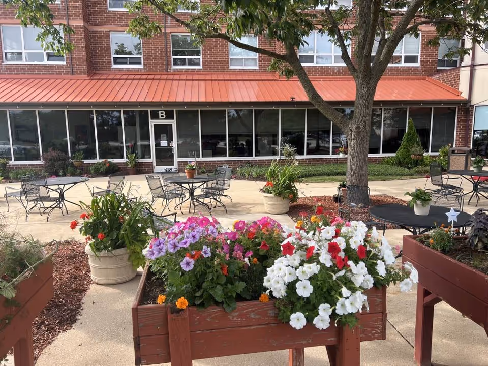 Outdoor patio area at Elison Independent Living of Niles with metal tables and chairs, surrounded by flower planters filled with colorful flowers including white, red, and purple blooms. A large tree provides shade, and a brick building with large windows and a red metal roof is in the background.