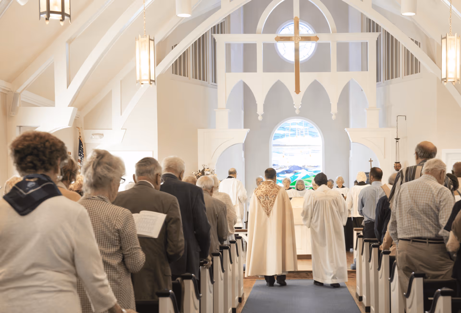 Interior view of a church or chapel during a religious service with elderly attendees standing in pews facing the altar where clergy members in robes are conducting the service. The space features high vaulted ceilings, hanging lights, and a large stained glass window behind the altar.