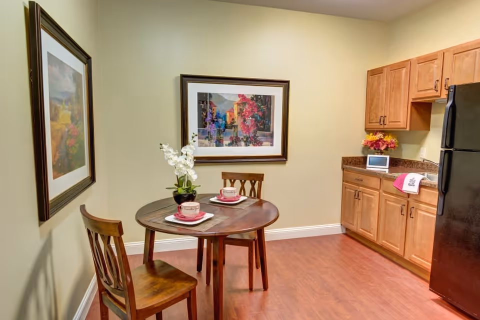 A small kitchen and dining area with wooden cabinets, a black refrigerator, and a round wooden table set for two with cups and plates. The walls are light-colored and decorated with two framed paintings. There is a small plant with white flowers on the table and a colorful flower arrangement on the countertop.