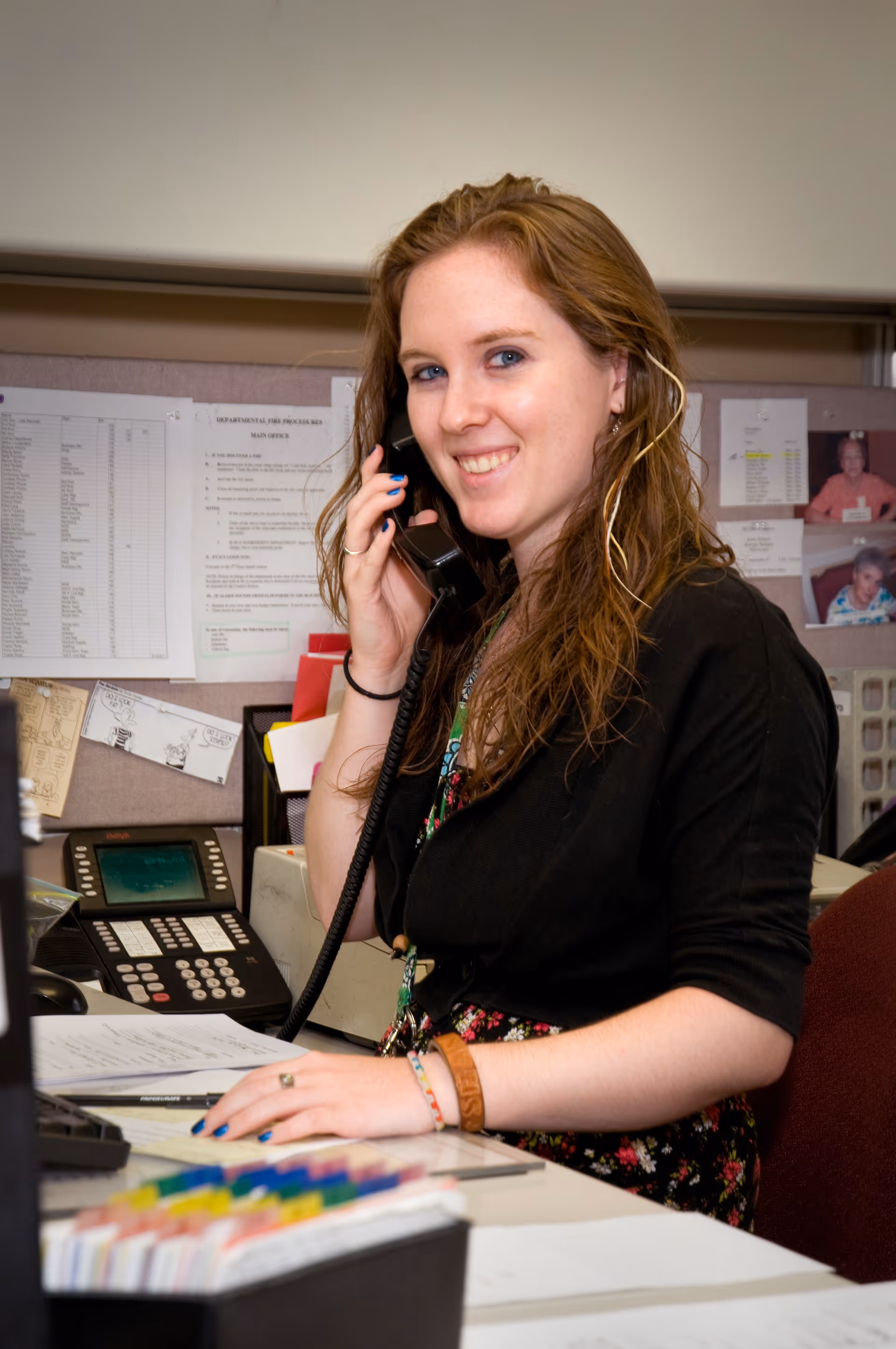 A young woman with long brown hair is sitting at a desk in an office environment, smiling while talking on a corded telephone. Behind her are pinned papers and photos on a bulletin board. The desk has office supplies, documents, and a multi-line phone system.