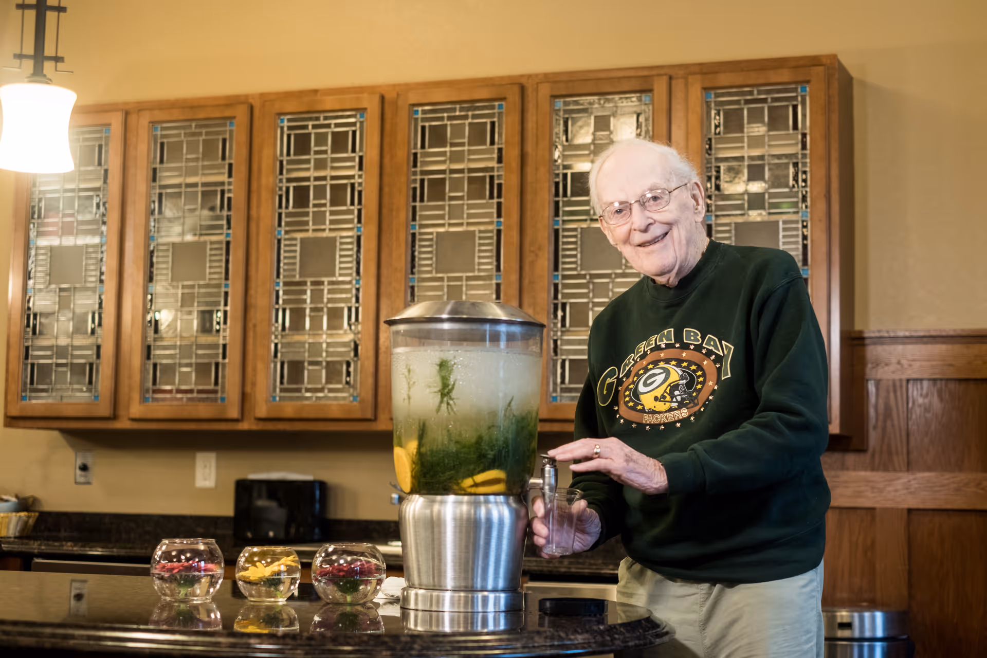 An elderly man smiles as he fills a glass from a beverage dispenser on a kitchen counter with cabinets behind him.