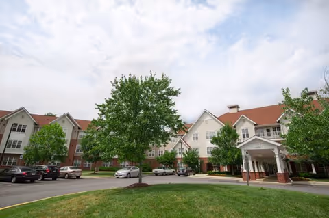 Exterior view of The Manor at Victoria Park, showing a large multi-story building with a red roof and white siding. There are several trees and parked cars in front of the building under a partly cloudy sky.