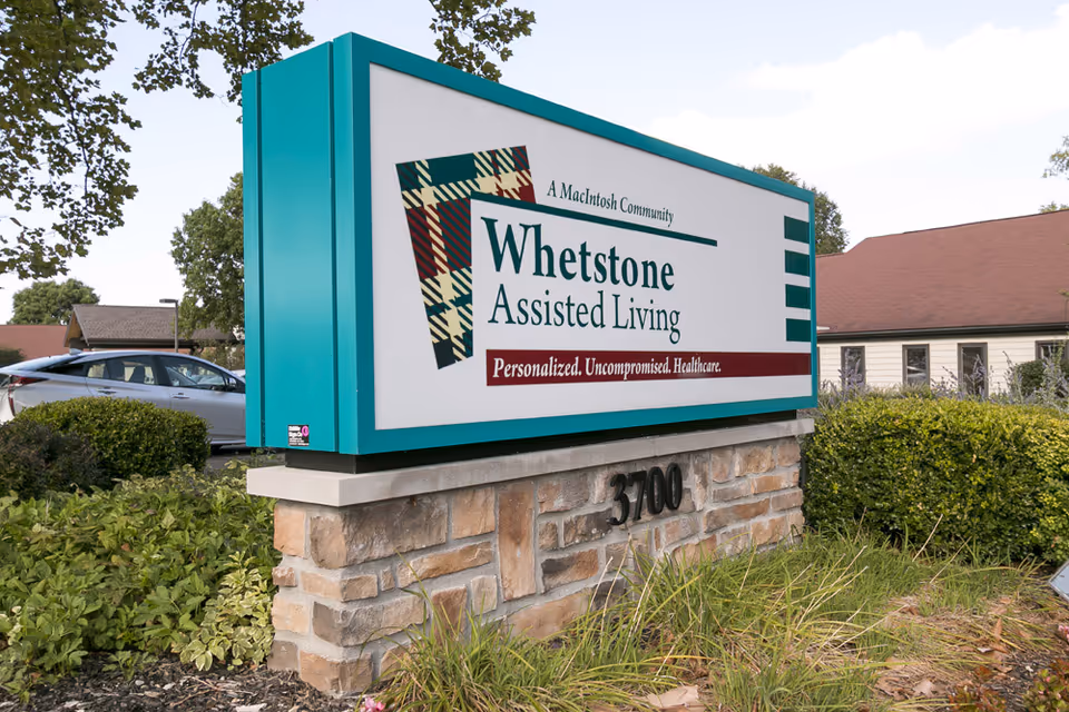 Outdoor sign reading "Whetstone Assisted Living" mounted on a stone base with landscaping and the facility building visible behind it.