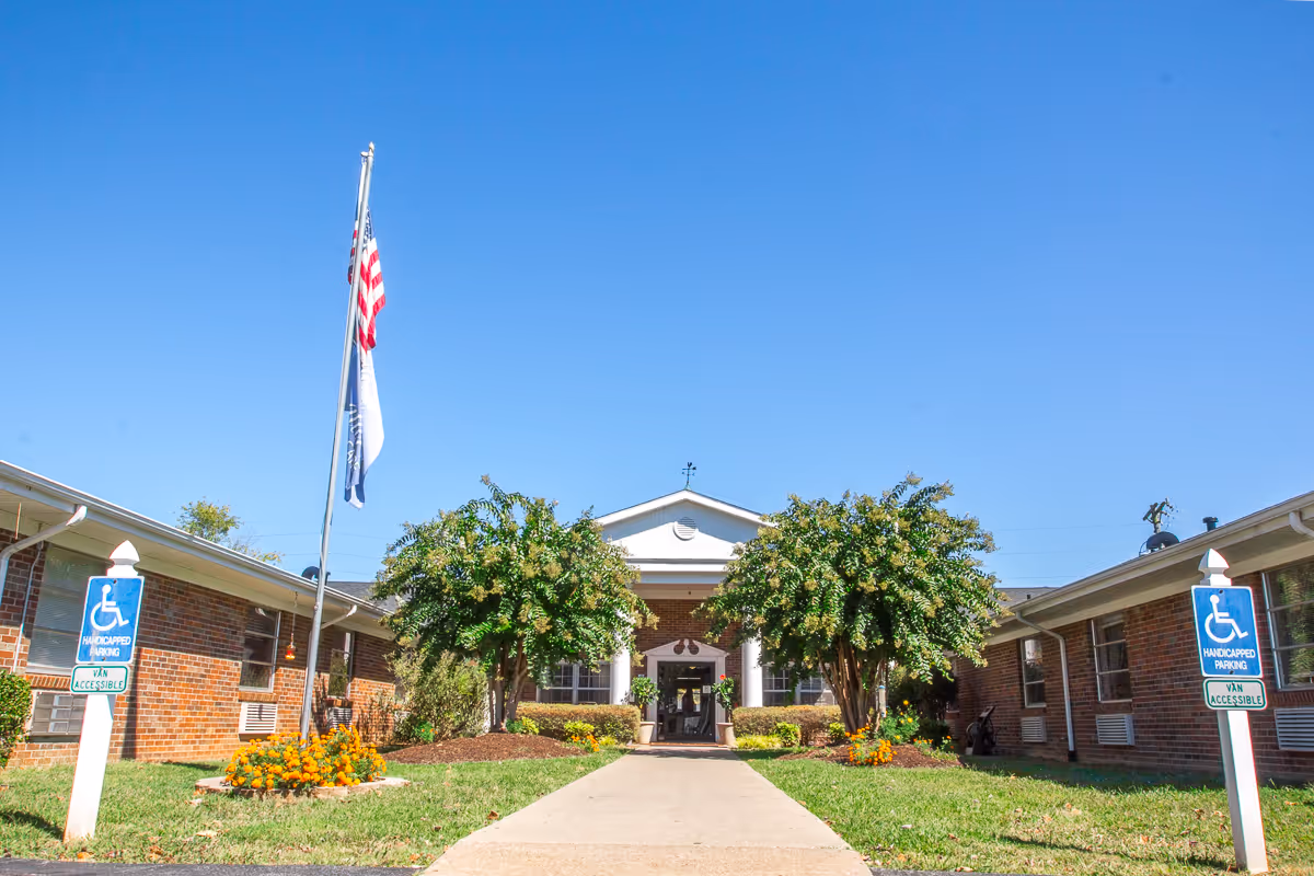 Front entrance of a single-story brick rehab and wellness center with a flagpole, walkway, landscaping, and handicap parking signs.