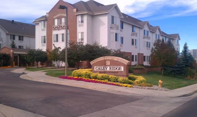 Exterior view of Caley Ridge Assisted Living facility showing a multi-story building with white siding and brick accents, surrounded by landscaped greenery and colorful flowers near the entrance sign.