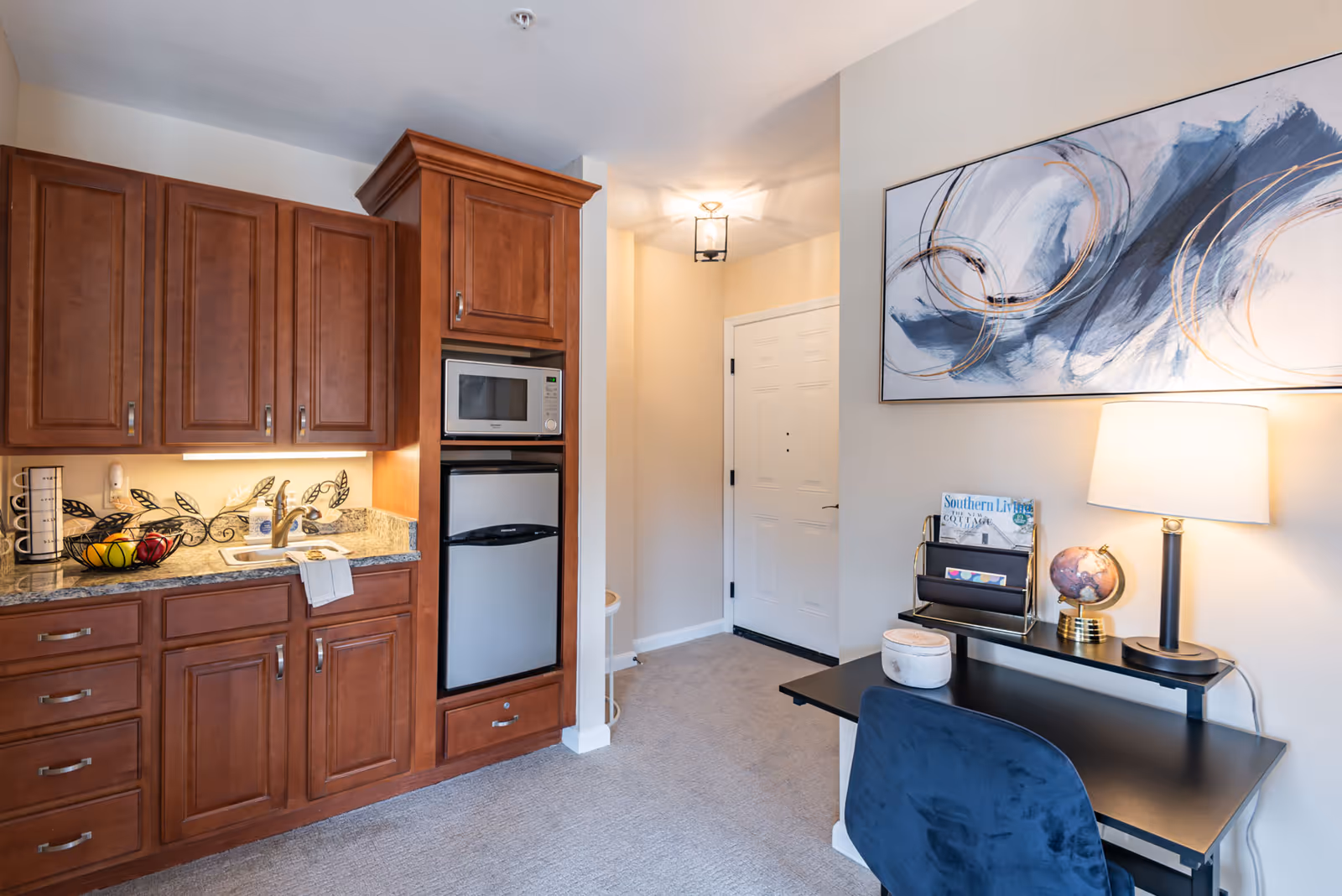 Interior view of a senior living facility room featuring a kitchenette with wooden cabinets, a microwave, and a mini refrigerator. Adjacent to the kitchenette is a small black desk with a blue chair, a table lamp, a globe, and a magazine holder. Above the desk is a large abstract painting with blue and gold swirls. The room has beige walls and carpeted flooring, with a white door and a ceiling light fixture in the background.