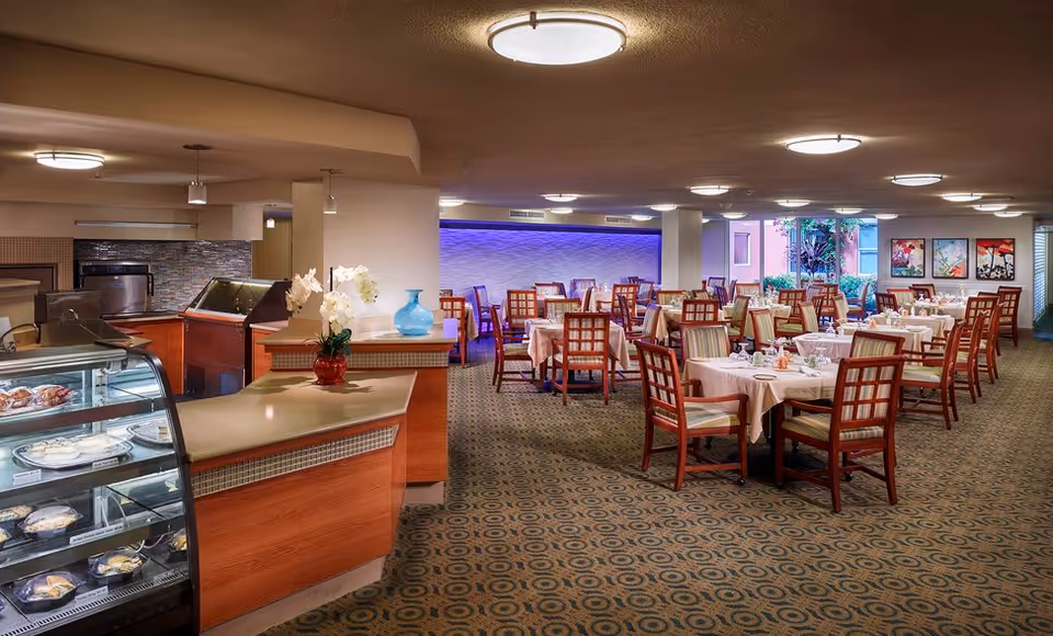 A spacious dining room with multiple tables covered in white tablecloths, each set with plates, glasses, and napkins. Wooden chairs with striped cushions surround the tables. On the left side, there is a counter with a glass display case containing desserts. The room is well-lit with ceiling lights, and there are decorative paintings on the far wall near a window showing some greenery outside.