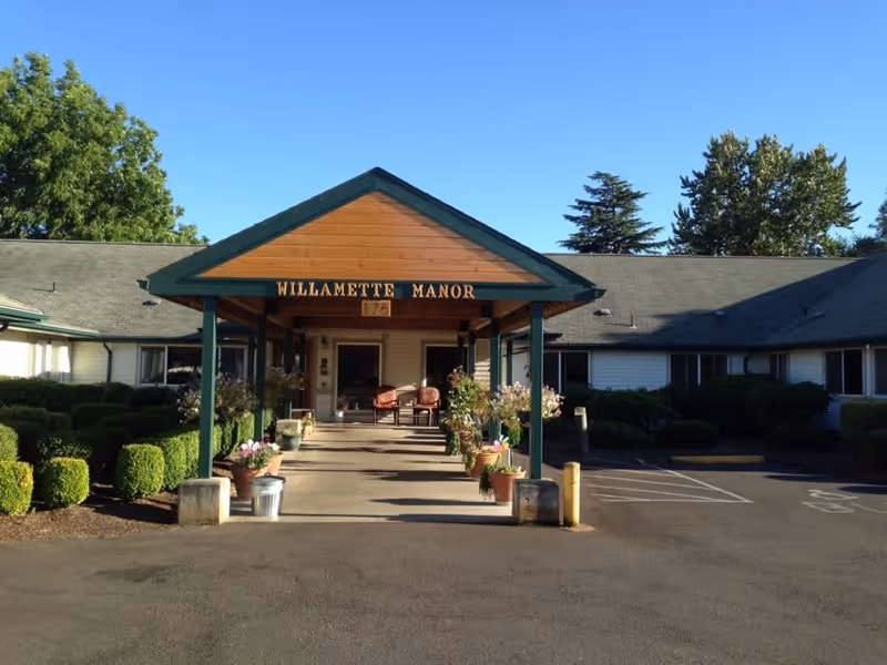 Front exterior view of Willamette Manor building with a covered entrance supported by green pillars, potted plants lining the walkway, and a clear blue sky above.
