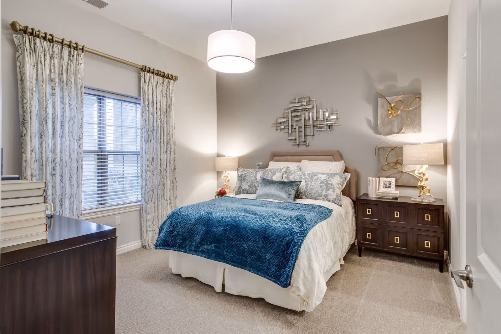 A neatly arranged bedroom with a bed covered in white and blue bedding, two bedside tables with lamps, decorative wall art above the bed, a window with patterned curtains, and a dark wooden dresser with a stack of books.