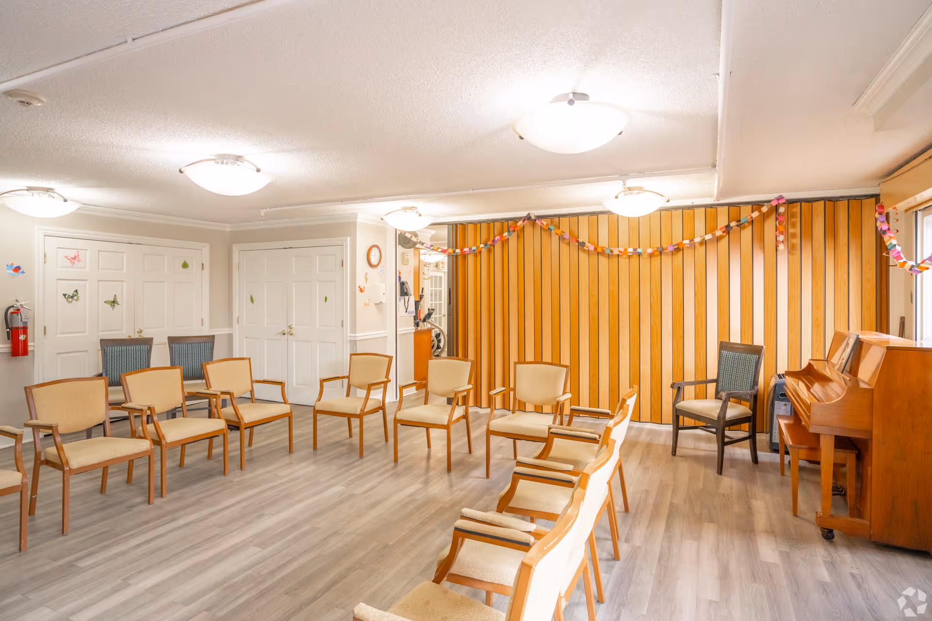 A bright room with wooden panel walls and light-colored flooring, arranged with multiple wooden chairs in a semi-circle. There is a piano on the right side and colorful paper chain decorations hanging on the wall. The room has several ceiling lights and white double doors on the left side.