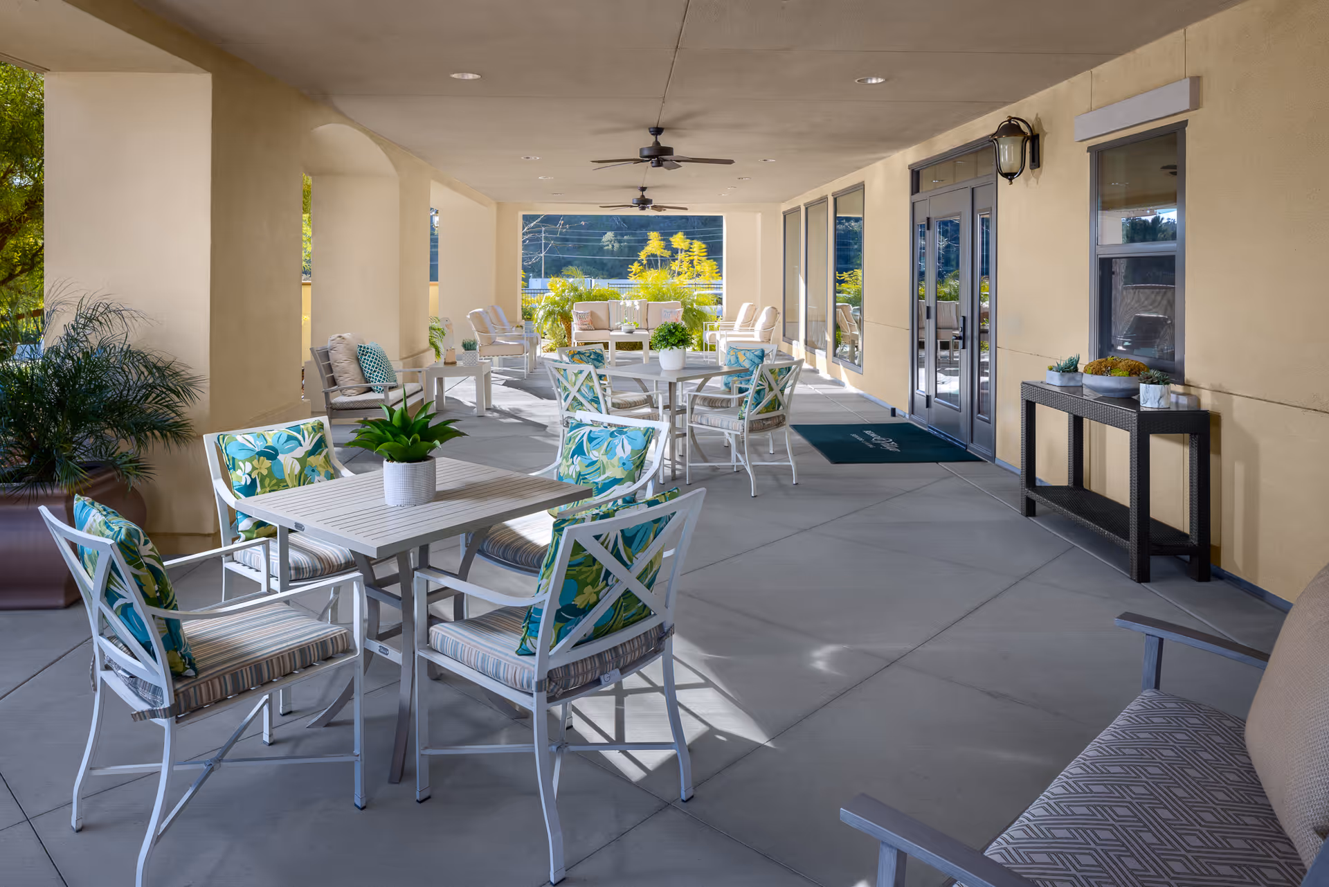 Covered outdoor patio area with multiple seating arrangements including tables and chairs with floral cushions, potted plants on tables, ceiling fans, and large windows and doors leading inside the building.