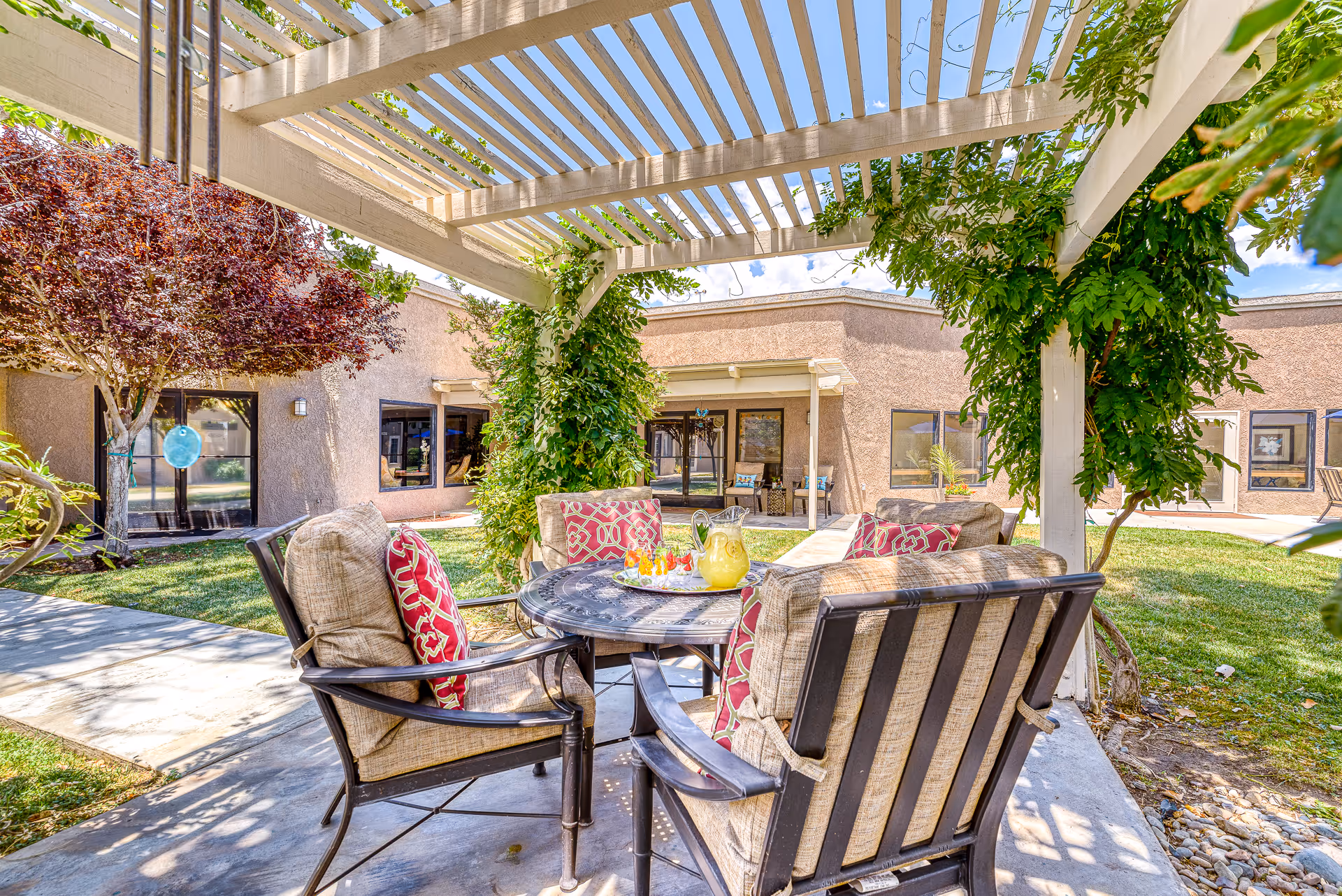 Shaded outdoor courtyard patio with cushioned chairs and a round table under a pergola in front of the facility.