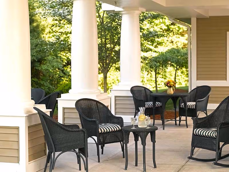 Outdoor covered patio area with black wicker chairs featuring striped cushions, small tables, white columns, and greenery in the background.