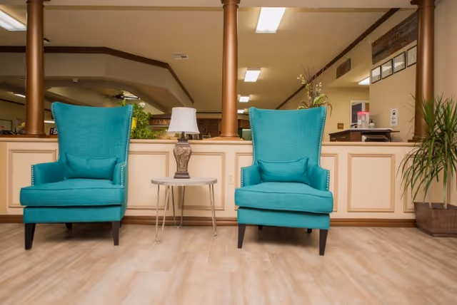 Two teal upholstered armchairs with matching cushions are positioned on either side of a small round table with a decorative lamp on it. The setting appears to be an indoor common area with beige walls, wooden columns, and potted plants in the background.