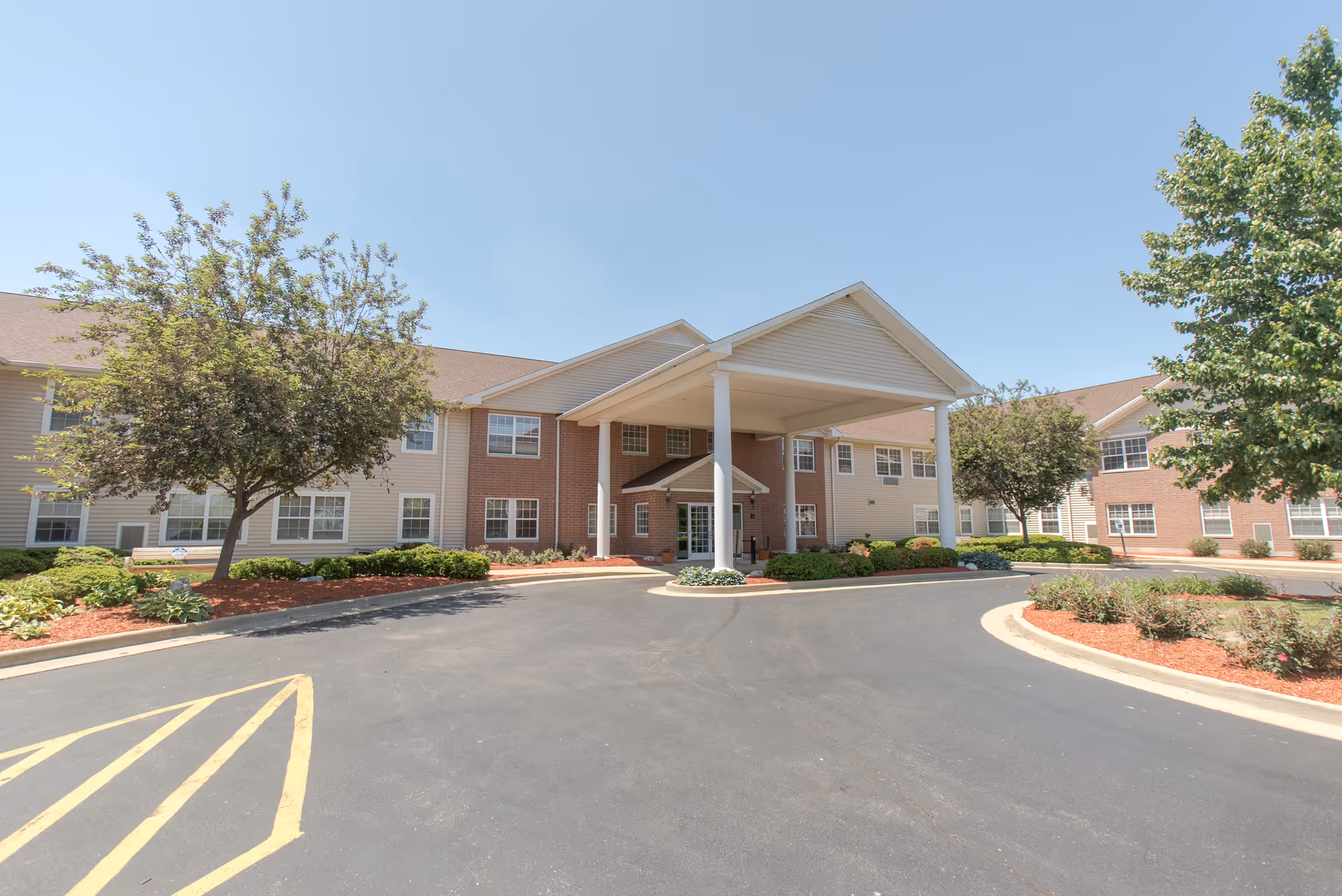 Exterior view of Bowman Estates of Danville, showing a two-story building with a covered entrance, surrounded by landscaped greenery and trees under a clear blue sky.