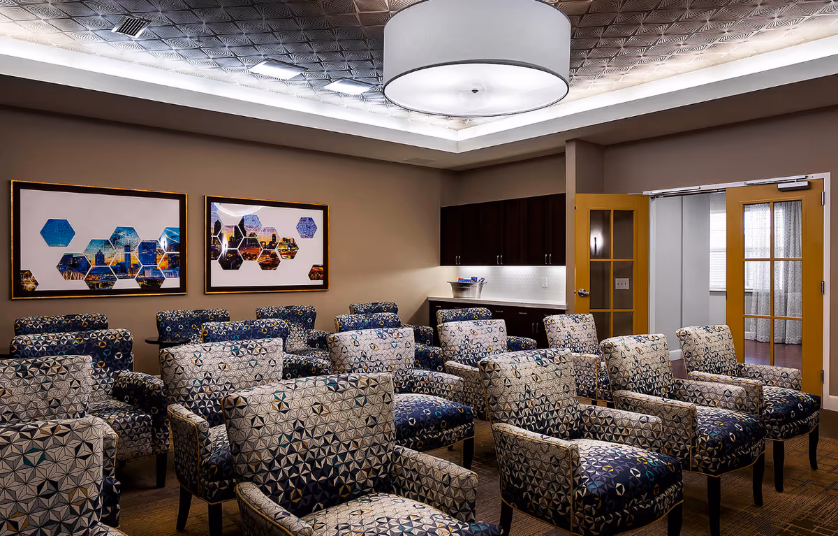 Well-lit common room with rows of patterned upholstered chairs facing forward, framed wall art, a kitchenette, and double doors.