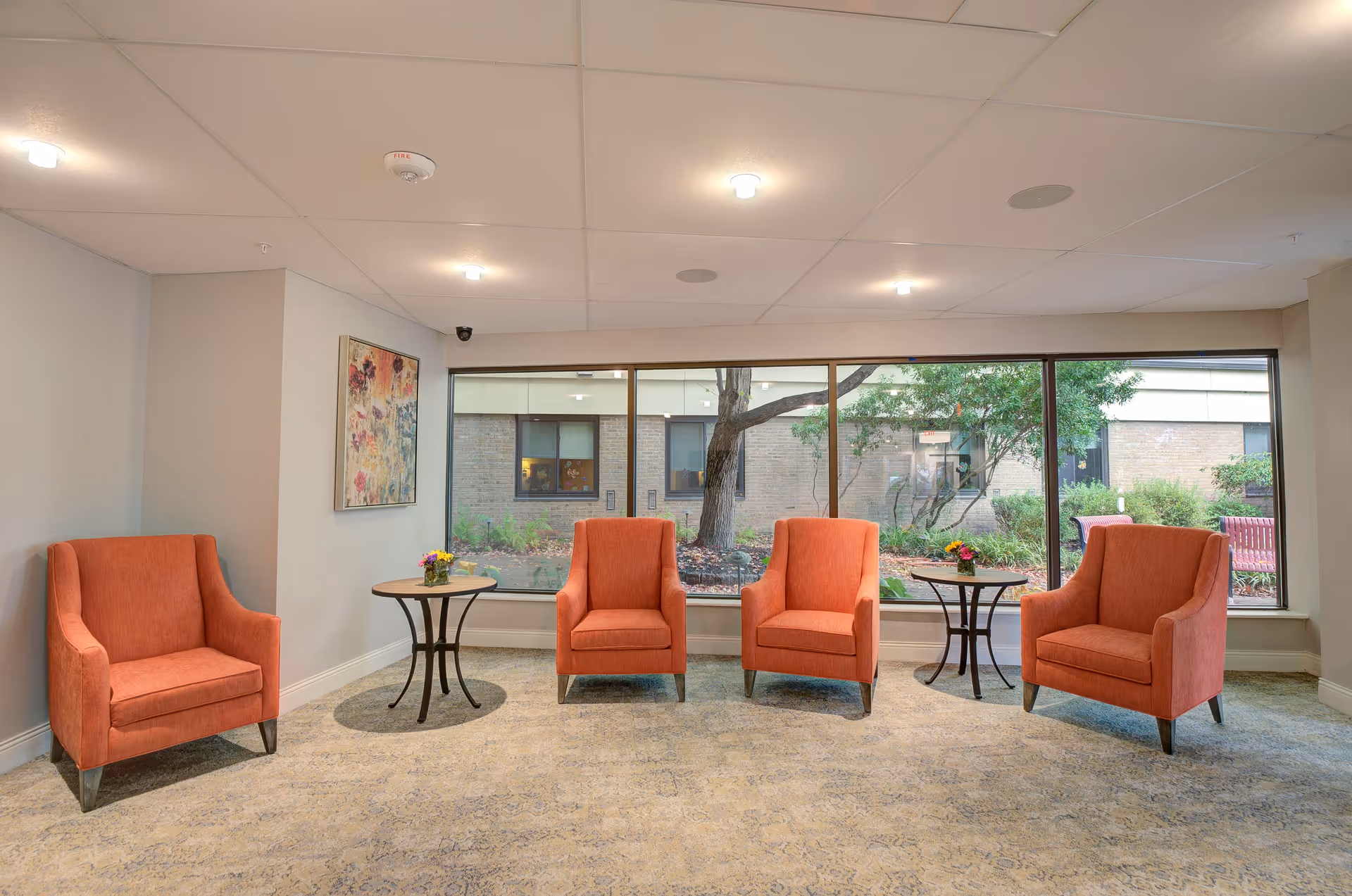 A bright seating area with four orange armchairs arranged in a semi-circle facing large windows that look out onto a garden with trees and shrubs. Two small round tables with flower arrangements are placed between the chairs. The room has a light-colored carpet and neutral walls with a colorful abstract painting on one wall.