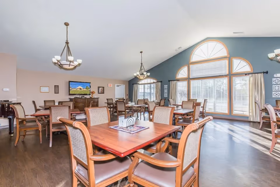 A bright and spacious dining room with multiple wooden tables and cushioned chairs arranged neatly. Large windows with arched tops allow natural light to fill the room. The walls are painted in soft beige and blue tones. Two chandeliers hang from the ceiling, and a television is mounted on the far wall above a wooden cabinet.