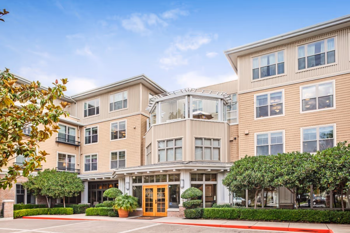 Front exterior of a multi-story assisted living building with a central glass entryway, potted plants, and trimmed landscaping.
