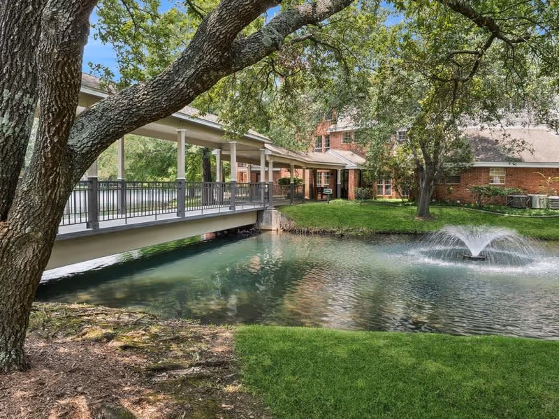 A peaceful outdoor scene at The Forum at The Woodlands featuring a small pond with a water fountain, a covered pedestrian bridge with railings, large trees providing shade, and a brick building in the background.